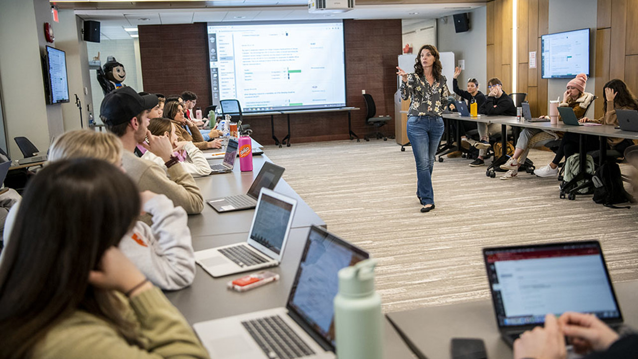 Lecturer in middle of room speaking to students who have open laptops
